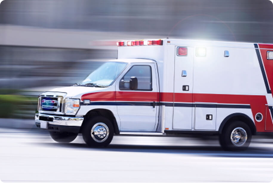A white and red ambulance speeds down a city street with lights flashing, conveying urgency and emergency response. The background is motion-blurred.