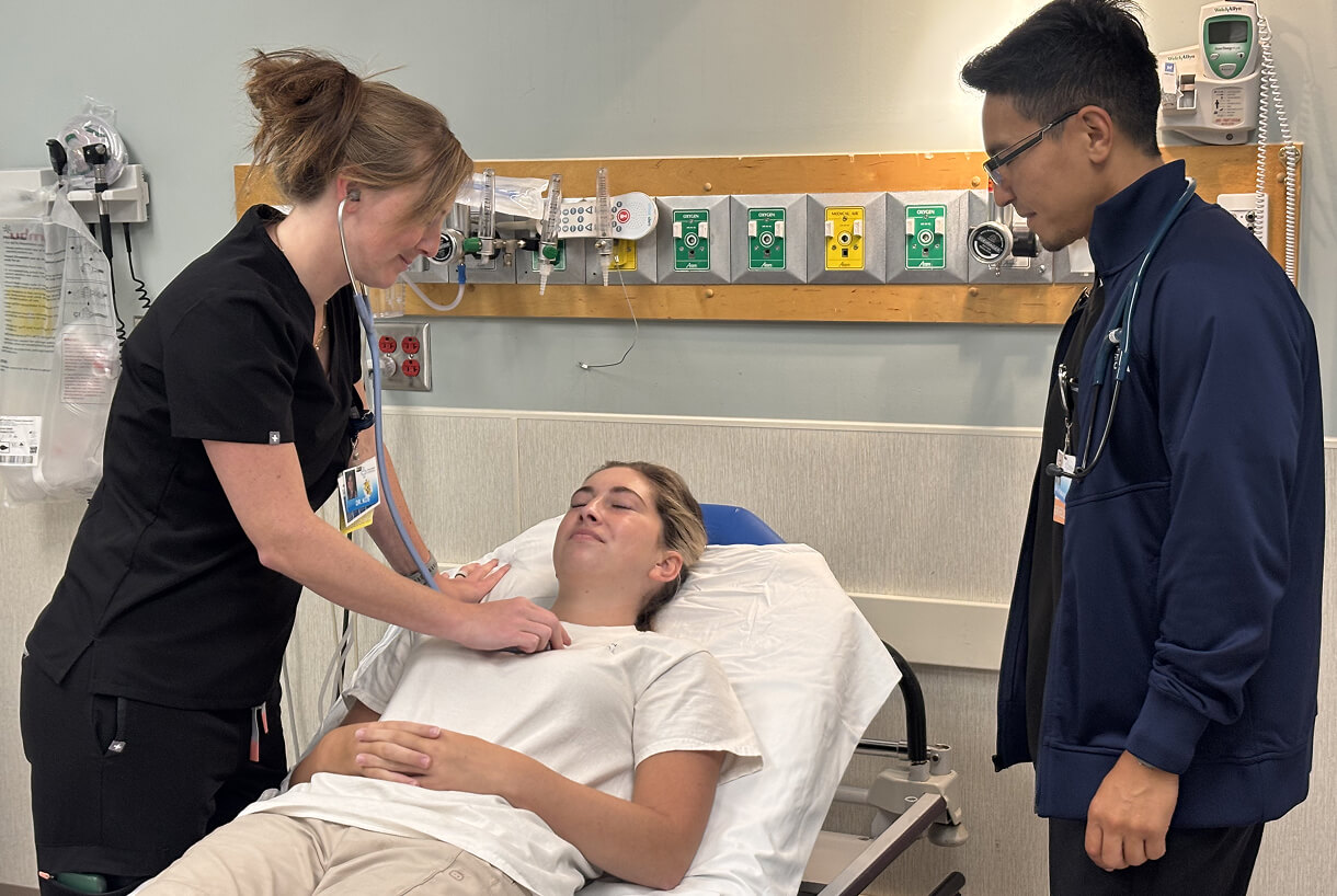 A nurse listens to a patient's chest with a stethoscope in a hospital room, while a doctor looks on. The atmosphere is calm and professional.